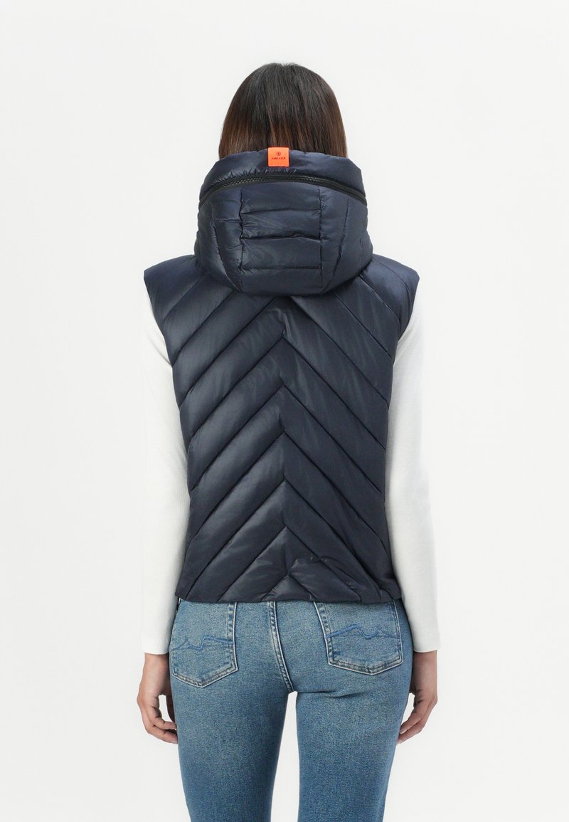 Navy-Wattweste mit Chevron-Muster und abnehmbarer Kapuze, mit einem orangefarbenen Etikett auf der Rückseite, getragen über einem weißen Langarmoberteil.