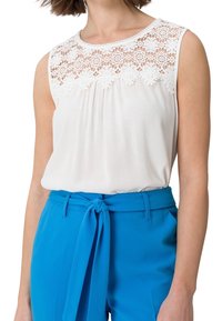 Top blanco sin mangas con detalles de encaje en los hombros, combinado con pantalones azules de tiro alto que cuentan con un cinturón atado.