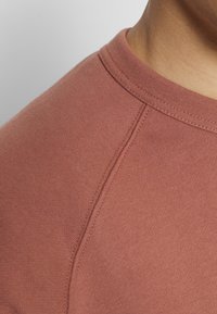 Camiseta de algodón en un color rosa apagado con cuello redondo, costuras visibles y una textura suave. Primer plano de la tela.