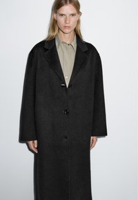 Cappotto di lana nera con una vestibilità rilassata, ampi revers e tre bottoni. Il tessuto appare morbido con una consistenza liscia, completato da una camicia verde chiaro sotto.