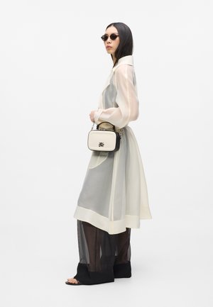 Femme portant un long manteau crème transparent sur un pantalon noir transparent, tenant un petit sac à main crème et noir, et portant des lunettes de soleil.