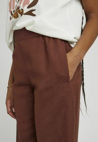 Pantalons marron, à jambes larges, avec une taille élastique et des poches latérales. Le tissu semble texturé et léger. Fait partie d'une tenue avec un haut graphique.