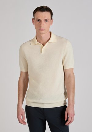 Mann in einem kurzärmligen cremefarbenen Poloshirt und schwarzer Hose, stehend vor einem schlichten hellgrauen Hintergrund.