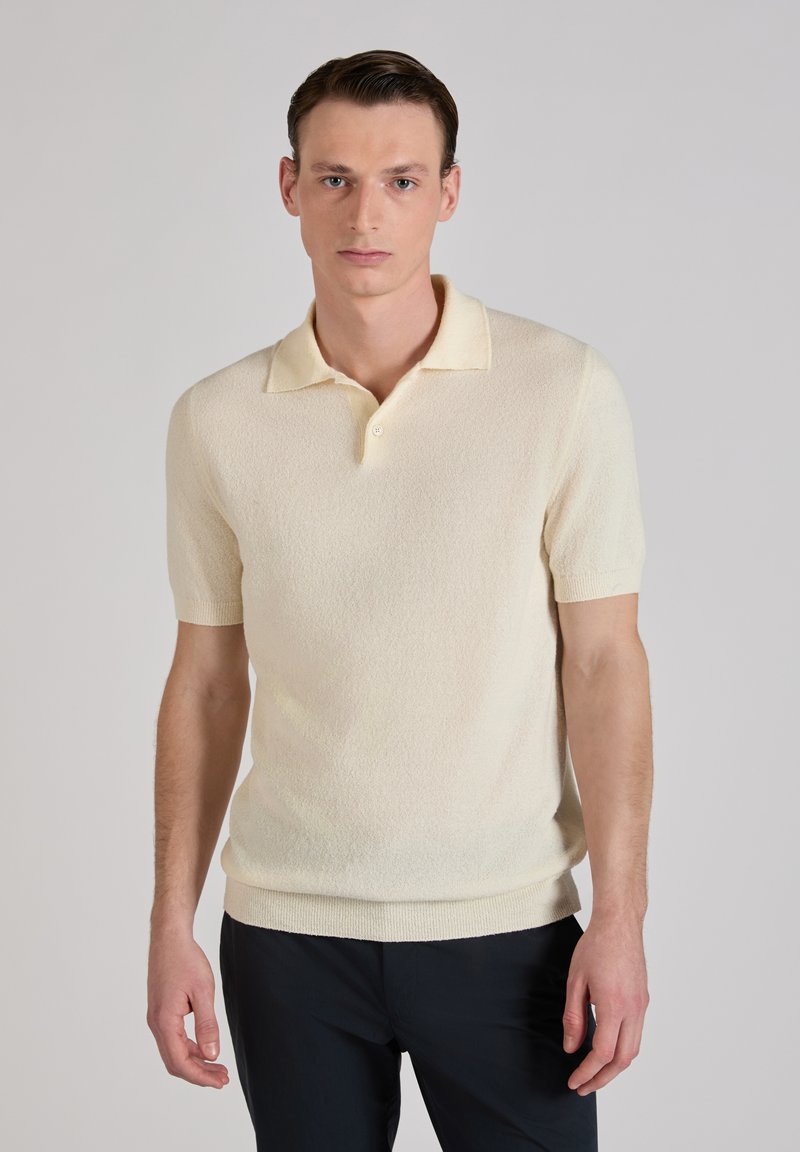 Mężczyzna ubrany w kremową koszulkę polo z krótkim rękawem i czarne spodnie, stojący na tle jednolitego jasnoszarego tła.