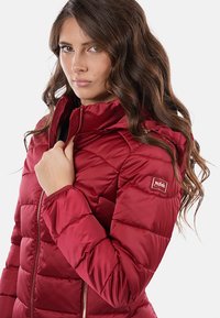 Giacca a vento rossa con colletto alto, design trapuntato, chiusura con zip e una toppa con logo sulla spalla. Tessuto liscio e lucido.