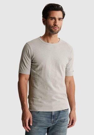 Mand med mørkt hår og skæg iført en lysebeige kortærmet striktrøje og blå jeans, stående mod en ensfarvet baggrund.