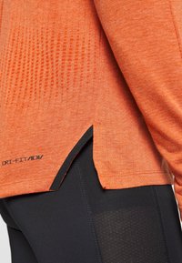 Pomarańczowa sportowa koszulka z długim rękawem o teksturowanej powierzchni, z bocznymi rozcięciami i czarnym akcentem. Na dole znajduje się logo Dri-FIT ADV.