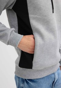 Sudadera gris con panel lateral negro, puños acanalados y un bolsillo. Tejido suave con textura lisa. Detalle de bolsillo con cremallera en el lateral.