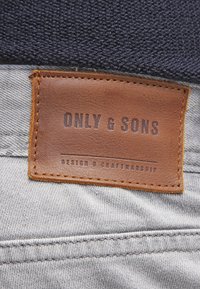 Ruskea nahkatarra, jossa kohokuvioitu teksti "ONLY & SONS" ja "DESIGN & CRAFTMANSHIP," kiinnitetty harmaaseen kangasmateriaaliin, jonka pinta on teksturoidut ja siinä on ompelut.