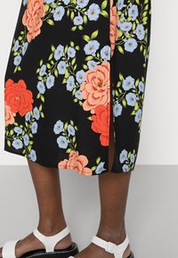 Vestido floral com grandes flores vermelhas e azuis claras sobre um fundo preto. Detalhe de fenda lateral. Sandálias brancas com tiras ajustáveis no tornozelo.