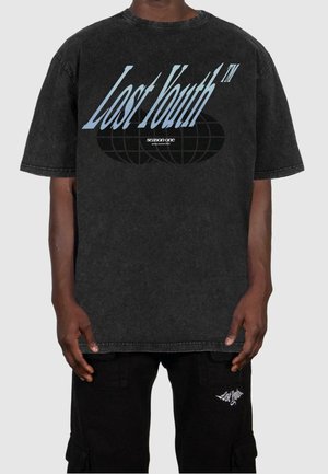Persona vistiendo una camiseta negra de gran tamaño con el texto "Lost Youth™" y un gráfico de un globo terráqueo, combinada con pantalones negros que tienen un pequeño logo blanco.