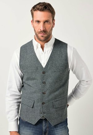 Gilet grigio in lana con scollo a V, cinque bottoni marroni, due tasche frontali, abbinato a una camicia bianca e jeans blu. Tessuto liscio, vestibilità sartoriale.