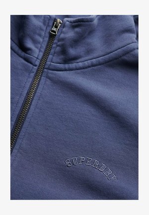 Zbliżenie niebieskiej bluzy rozpinanej na zamek z metalowym zamkiem błyskawicznym oraz haftem "Superdry" w dopasowanym niebieskim kolorze na piersi.