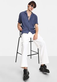 Camisa de manga corta azul marino con patrón texturizado, combinada con pantalones anchos blancos y zapatos negros gruesos, sentado en un taburete negro.