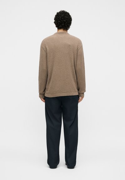 Άτομο με σγουρά μαλλιά στέκεται με την πλάτη προς την κάμερα, φοράει καφέ υφασμάτινο μακρυμάνικο τοπ και φαρδιά μαύρη παντελόνα σε απλό φόντο.