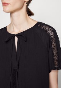Top nero realizzato in tessuto leggero, con chiusura a legatura al collo, dettagli in pizzo sulle spalle e delicata orlatura a zig zag.