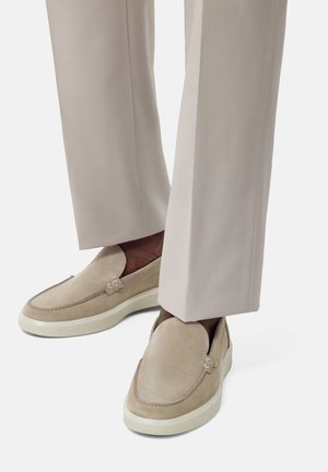 Beige Wildleder-Loafer mit weißen Gummisohlen, die mit Steppnähten und einem Logodesign versehen sind, kombiniert mit hellbeigen, tailleurartigen Hosen.
