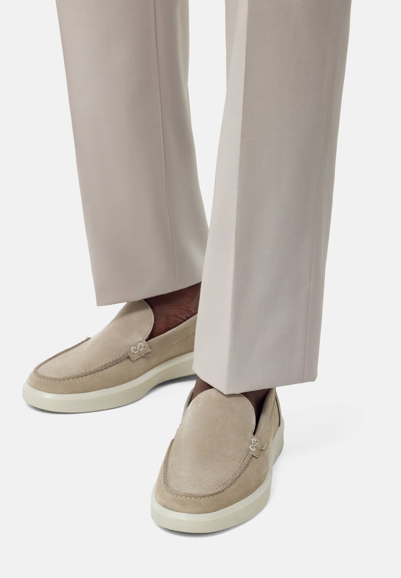 Beige suede loafers with white rubber soles, featuring stitching details and a logo accent, paired with light beige tailored trousers.