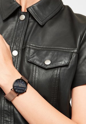 Chemise en cuir noir avec des boutons-pression et des poches poitrine. Une montre-bracelet avec un cadran noir rond et un bracelet en maille rose gold est affichée sur un poignet.