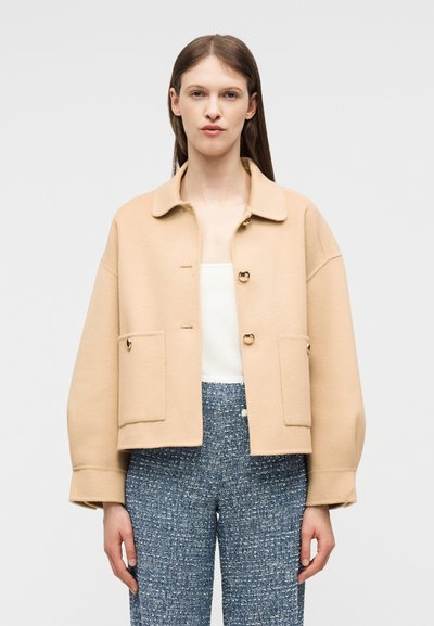 Γυναίκα που φοράει μπεζ σακάκι με κουμπιά και μεγάλες τσέπες, καθώς και μπλε παντελόνια με σχέδια, μπροστά σε ένα απλό φόντο.