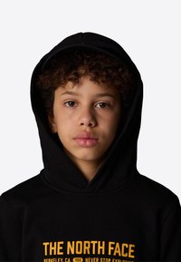Hoodie nero con tasca frontale a marsupio, cappuccio regolabile con coulisse e logo "THE NORTH FACE" in giallo sul petto.