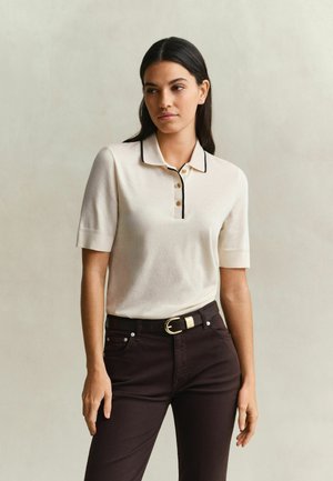 Femme debout, portant une chemise crème à manches courtes et col, avec des bordures et boutons noirs, un pantalon sombre et une ceinture noire avec une boucle dorée.