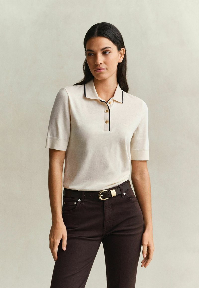 Femme debout, portant une chemise crème à manches courtes et col, avec des bordures et boutons noirs, un pantalon sombre et une ceinture noire avec une boucle dorée.