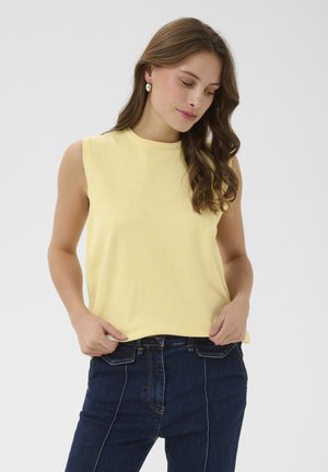 Hullámos barna hajú nő gyöngyfülbevalóval, ujjatlan sárga felsőben és sötétkék farmerral, zsebre tett kézzel áll.