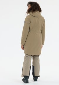 Khaki vandafvisende parka med hætte, bælte i taljen og sidelommer. Stylet med beige bukser med sorte detaljer og kraftige sorte støvler.