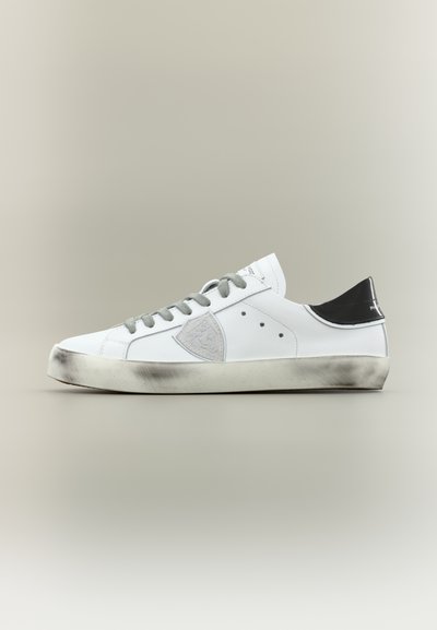 Baskets basses blanches avec lacets gris, une pièce latérale grise et une languette de talon noire, montrant les détails de la semelle usée sur un fond neutre.