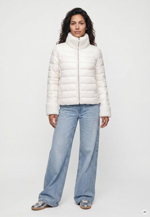 Frau, die nach vorne schaut, trägt eine weiße gepolsterte Jacke, hellblaue Jeans mit weitem Bein und silberne Sneakers vor einem schlichten Hintergrund.