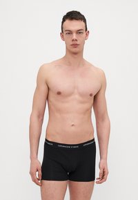 Svarte boxer-shorts med bred, merket midje. Glatt tekstur, tettsittende design og midt på låret lengde. Modellen har bar overkropp.