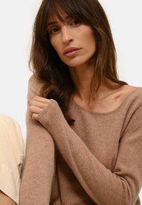 Hellbrauner Strickpullover mit weitem Ausschnitt und langen Ärmeln. Weiche Textur und lockere Passform, kombiniert mit einem leichten Stoff darunter.