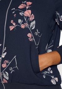 Giacca zip-up blu navy con stampa floreale in rosa e grigio, tessuto morbido, polsini a coste e tasche laterali. Texture liscia con vestibilità comoda.