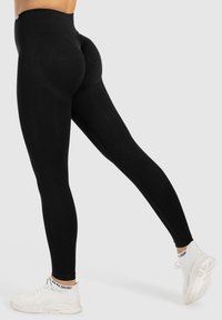 Leggings negros de cintura alta hechos de una tela elástica y suave. Presentan un diseño ajustado que resalta la figura, combinados con zapatillas blancas.