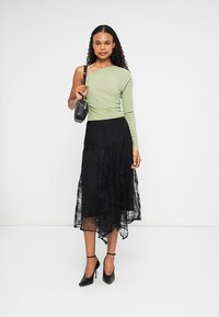 Top verde de um ombro só combinado com uma saia assimétrica de renda preta. O visual é complementar com sapatos de salto alto pretos e uma pequena mala.