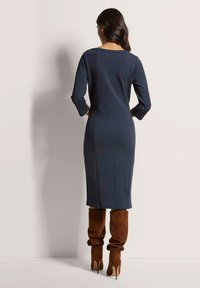 Navyfarbenes Langarmkleid mit taillierter Silhouette, gerippter Textur und Taillendetails, kombiniert mit braunen Wildleder-Stiefeln über dem Knie.