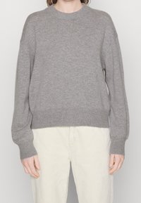 Person in einem lockeren grauen Strickpullover mit langen Ärmeln und cremefarbenen Cordhosen, der vor einem schlichten Hintergrund steht.