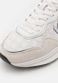 Gros plan de l’avant d’une basket blanche avec tissu texturé, accents en daim beige, lacets blancs et embout de semelle en caoutchouc noir.