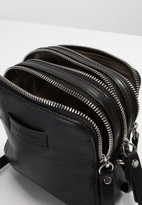 Czarna skórzana torba z trzema komorami zamykanymi na suwak, srebrnymi elementami i teksturowanym wykończeniem. Z boku znajduje się etykieta marki.