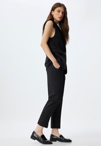 Blazer sans manches noir avec un bouton unique, associé à un pantalon noir sur mesure. Le look est complété par des mocassins noirs en cuir verni.
