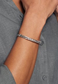 Srebrna pleciona metalowa bransoletka noszona na nadgarstku, z cylindrycznym zapięciem, noszona na szarej teksturowanej koszuli z detalami guzików.