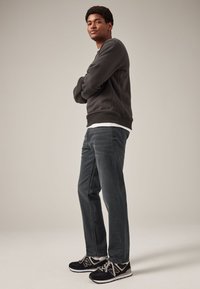 Jeune homme en sweat-shirt foncé, jean et baskets noires, debout les bras croisés, regardant légèrement vers le haut sur un fond uni.