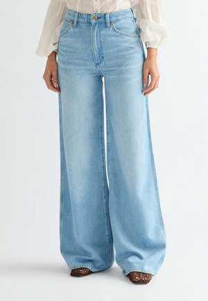 Kobieta ubrana w jasnoniebieskie, szerokie spodnie dżinsowe z wysokim stanem, zestawione z kremową bluzką z długim rękawem i brązowymi plecionymi butami.