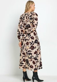 Blumen-Midi-Kleid aus weichem beigem Stoff mit dunkelroten, gelben und schwarzen Blumenmustern. Hat lange Ärmel und eine taillierte Silhouette. Getragen mit schwarzen Knöchelstiefeln.