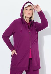 Longue veste zippée violette avec capuche, dotée d'accents roses et de poignets côtelés. Texture lisse, associée à un haut rose en dessous.