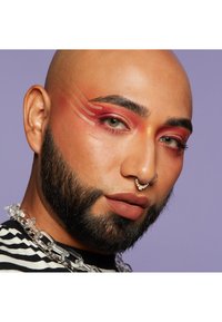 Skallig person med ett tjockt svart skägg, iförd en randig skjorta och ett klart kedjehalsband. Klara röda och orange ögonskuggor med slående mönster.