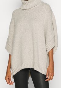 Helles beige Strickpullover mit Stehkragen, weiten Ärmeln und abgerundetem Saum. Strukturierte Wolle sorgt für subtile visuelle Akzente. Kombiniert mit dunklen Hosen.