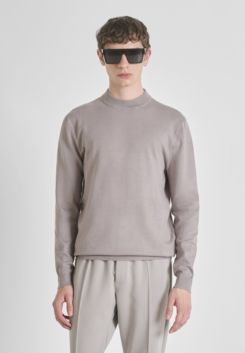 Giovane uomo che indossa occhiali da sole neri oversize, un maglione a maniche lunghe di un leggero color malva e pantaloni leggeri grigi coordinati, in piedi davanti a uno sfondo bianco.