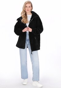 Czarna polarowa kurtka z kapturem, zapinana na zamek błyskawiczny, z opuszczonymi ramionami. Połączona z jasnoniebieskimi dżinsami i białymi trampkami.